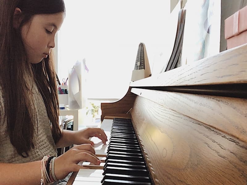 A child focuses on her hands while playing an acoustic piano during an online piano lesson