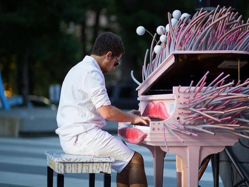 Man with sunglasses plays a grand piano with sculpted lips and bright pink and blue poles